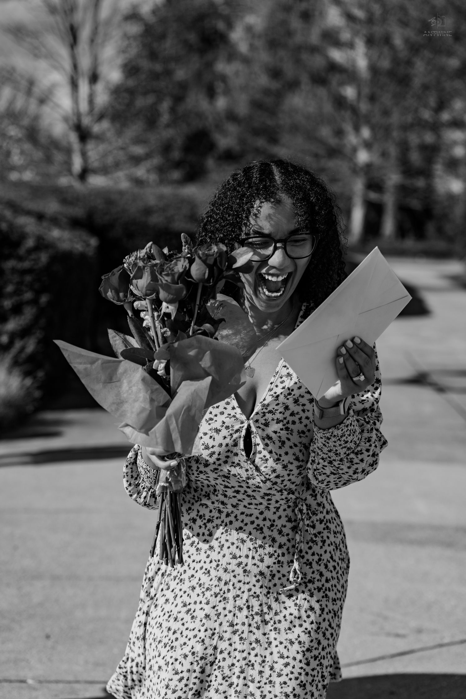 A happy woman with glasses reads a letter while holding a bouquet of flowers outdoors in black and white.