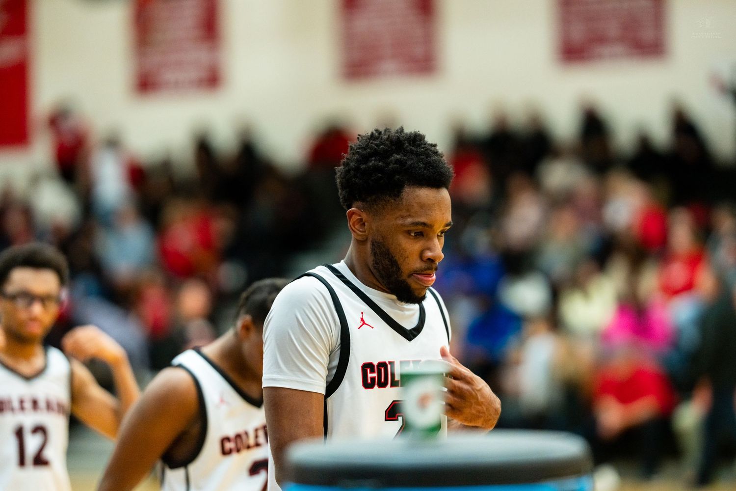 College basketball player in white Colgate jersey stands on court during a game with fans in background.