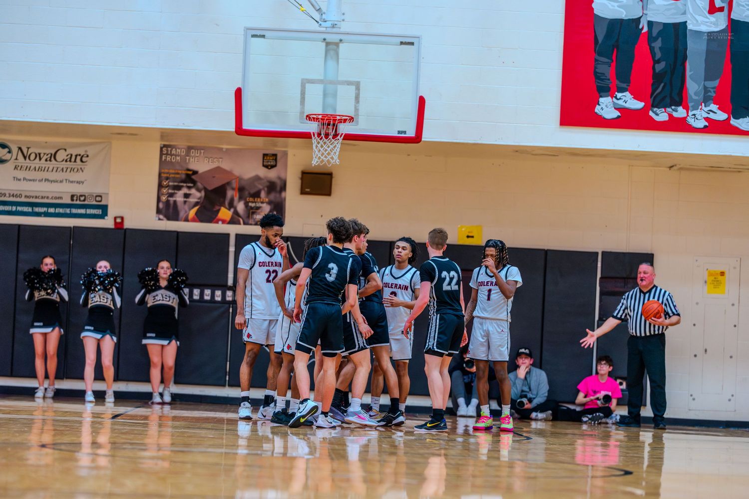 High school basketball players huddle near the basket during a timeout while cheerleaders watch from the sideline.