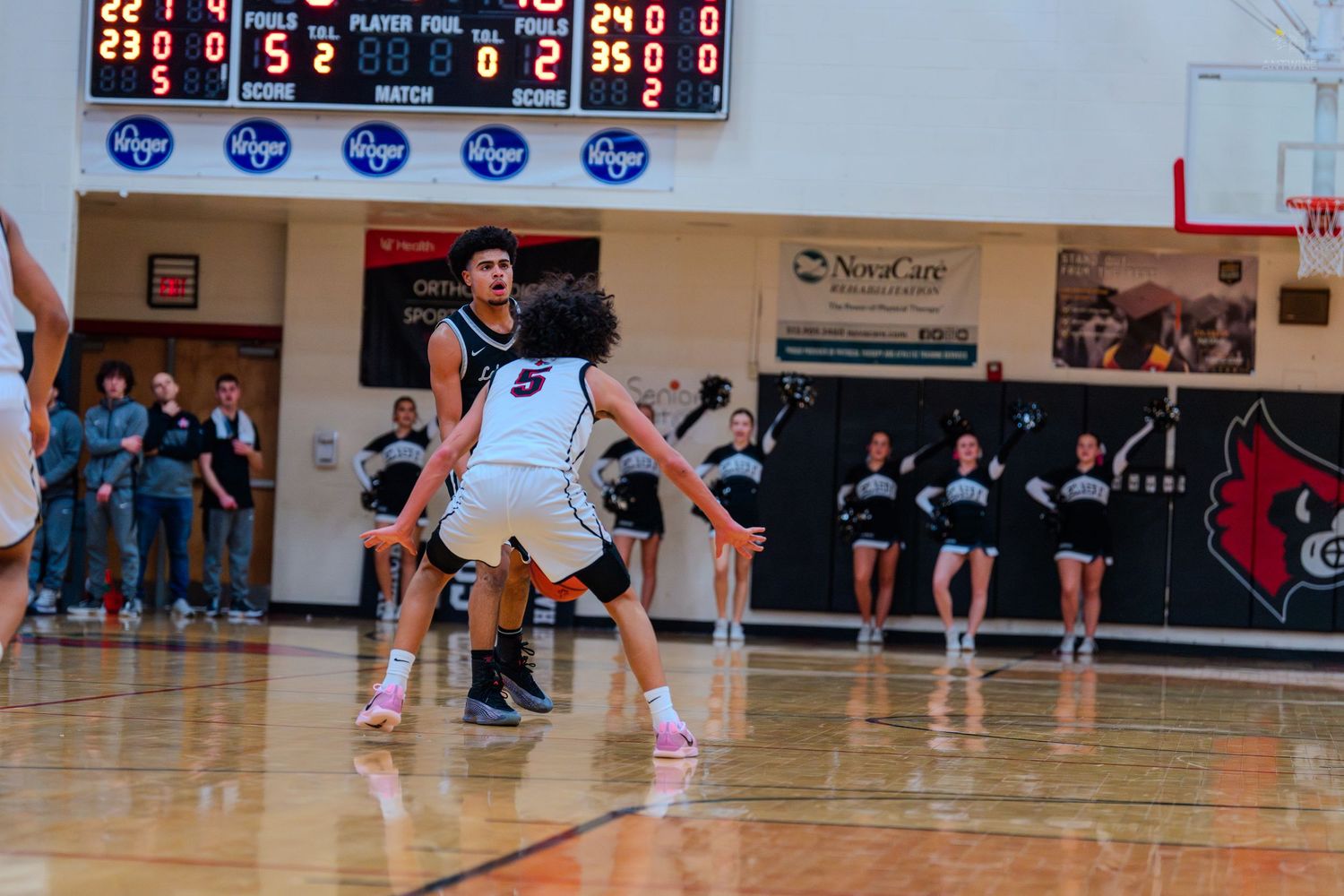 High school basketball player dribbles past a defender during an indoor game with cheerleaders visible in the background.