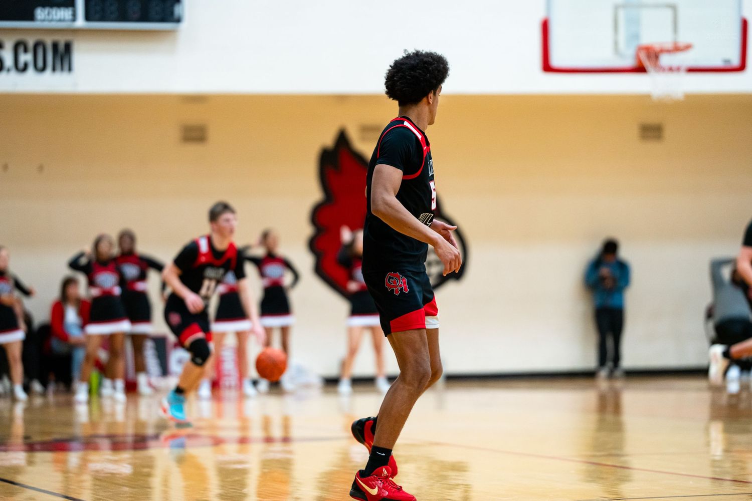 High school basketball player in black uniform dribbles ball near Cardinals cheerleaders on sideline.