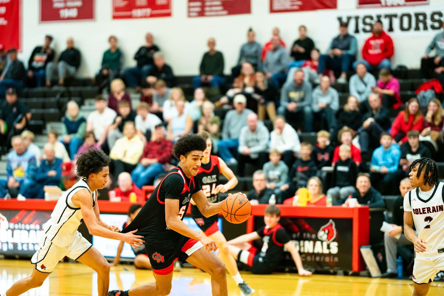Basketball player drives through defenders during competitive high school game in packed gym.