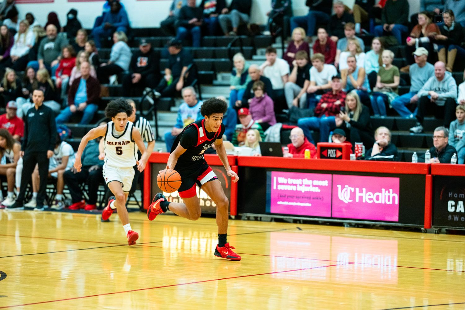 High school basketball player in red uniform drives past a defender during an intense game with fans in the background.
