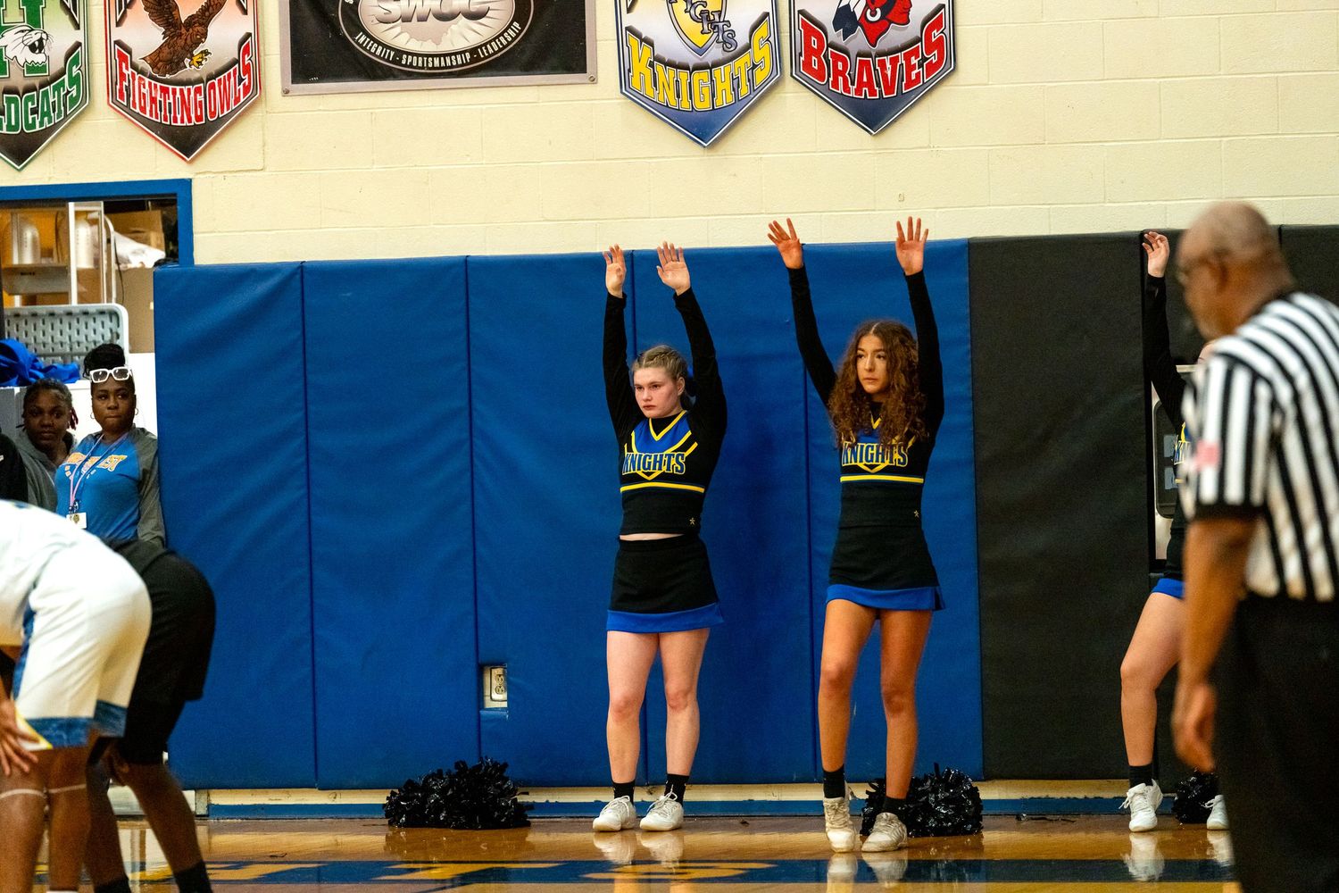 Two cheerleaders in blue and gold uniforms raise arms during a high school basketball game.