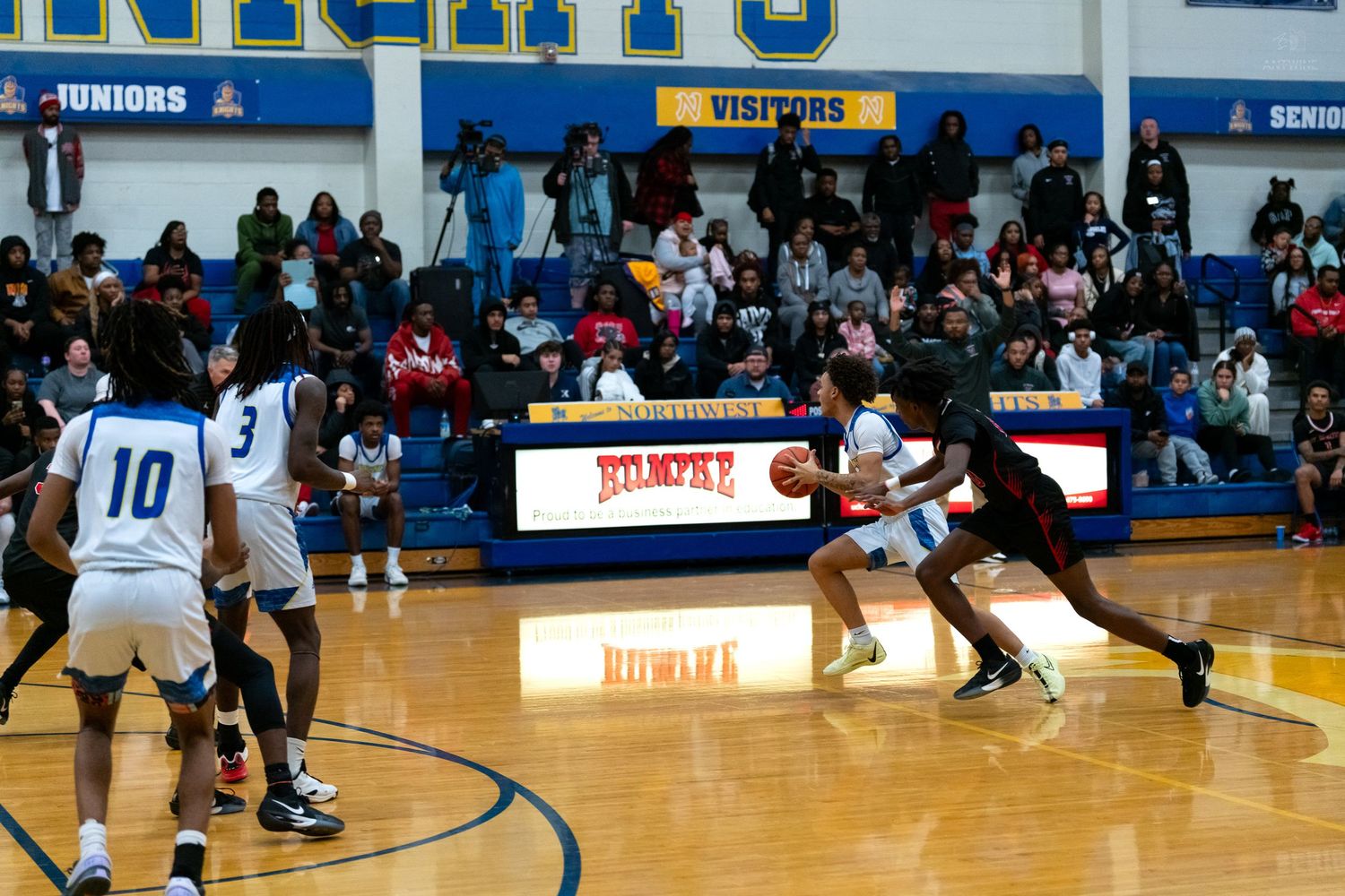 A basketball player drives toward the basket while defenders attempt to stop the play.
