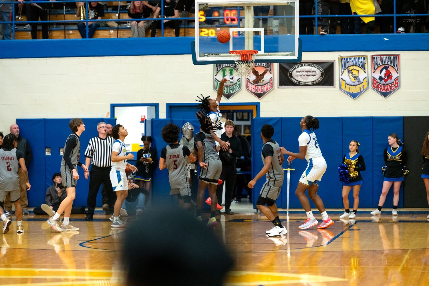 Players and referee gather under the basketball hoop during an intense high school gymnasium basketball game.