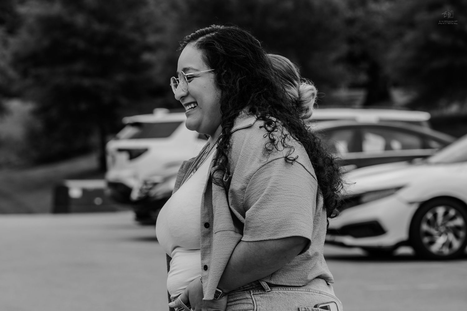 A woman with curly hair and glasses laughs joyfully in a parking lot in this black and white photo.