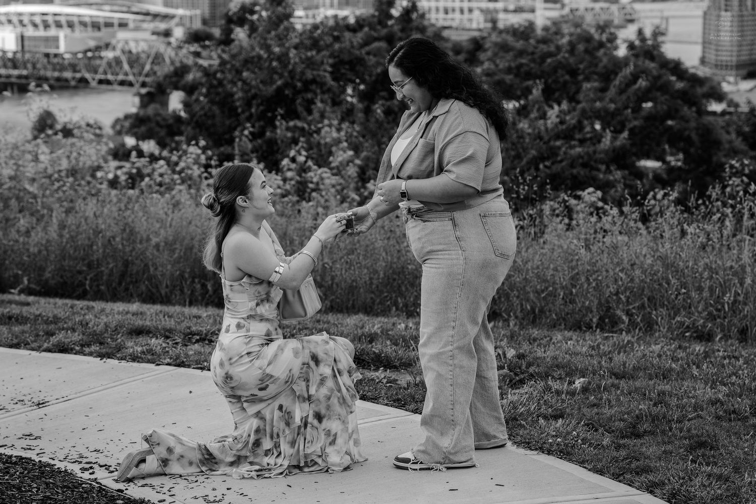 Woman kneeling on pathway proposing to partner outdoors with city skyline in black and white.