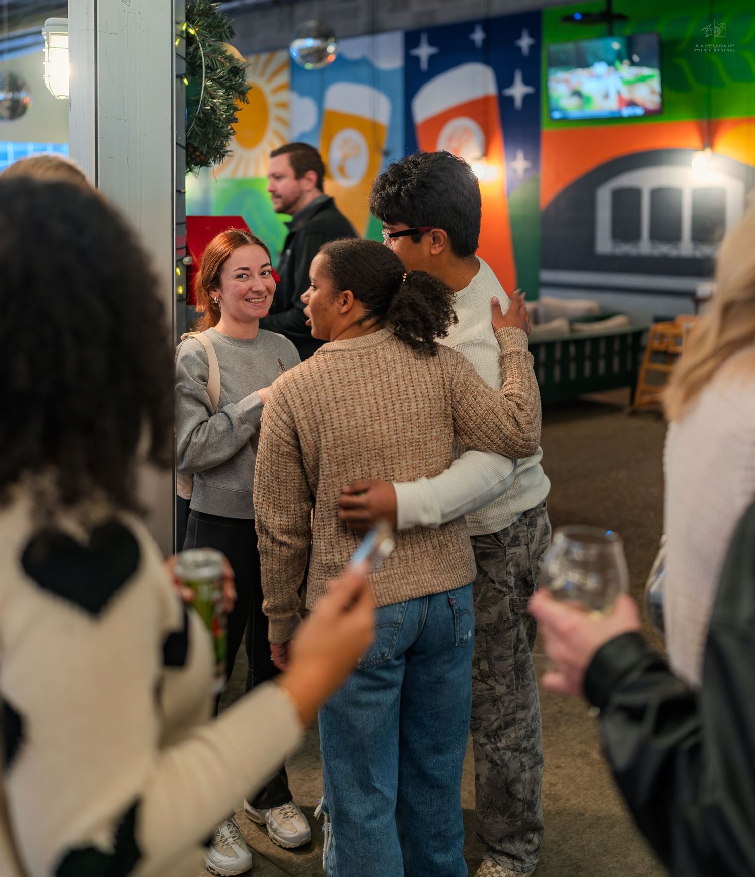 Friends hugging and socializing inside a colorful brewery venue during a casual gathering.