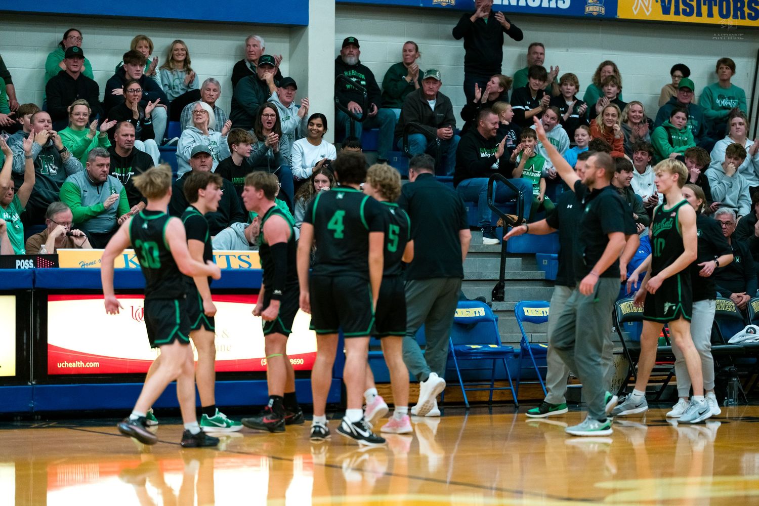 Green-uniformed basketball players celebrate on the court during a high school game with a packed crowd in the background.