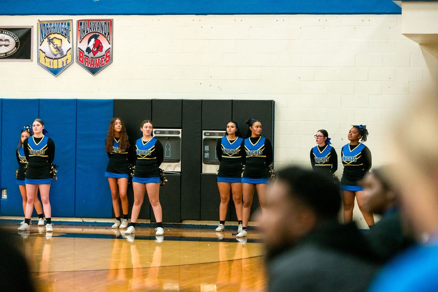 High school cheerleaders in blue and black uniforms stand along gymnasium wall watching basketball game from the sideline.