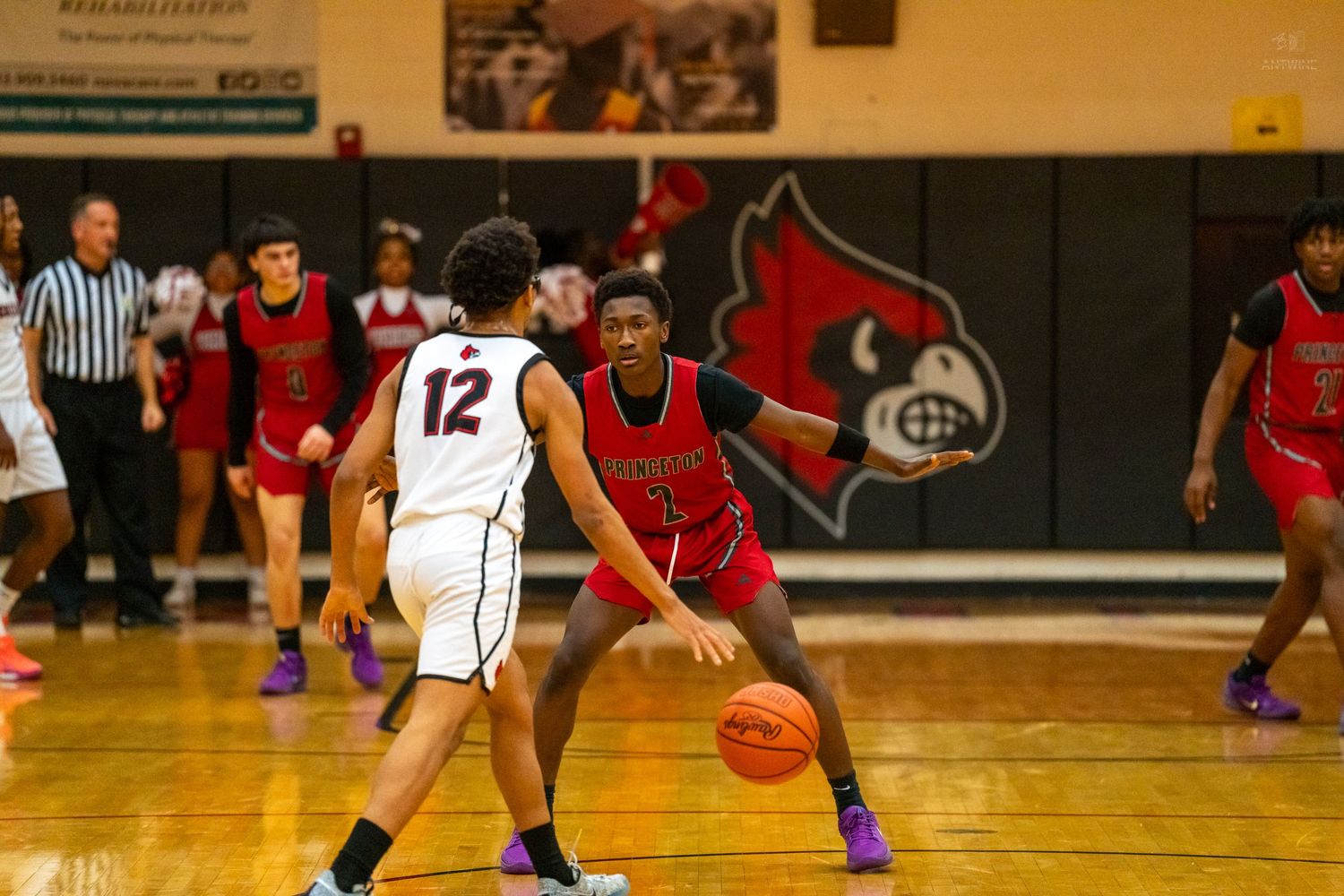 Basketball player #12 dribbles while defender in red uniform guards closely.