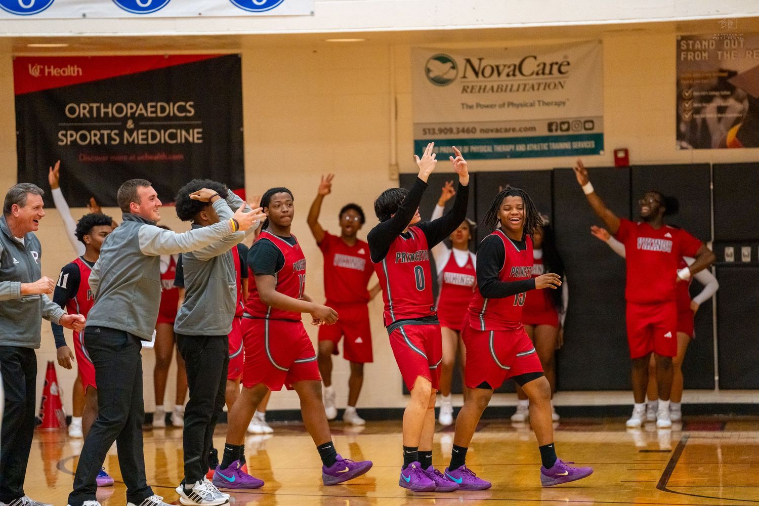 A high school basketball team in red uniforms celebrates enthusiastically on the sideline with coaches.
