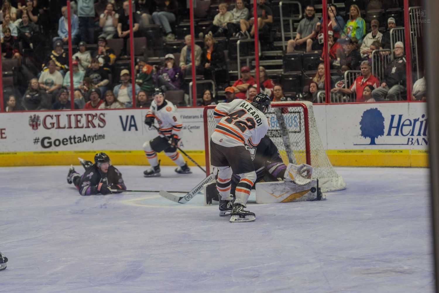Orange-jersied hockey player celebrates near net as opposing goalie and defender fall to ice after goal scored.