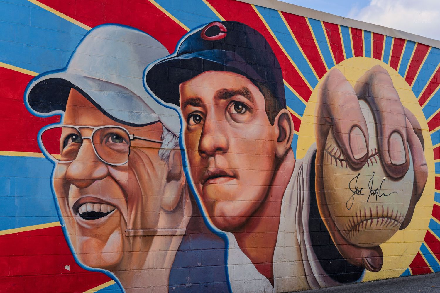 Colorful baseball-themed mural featuring two men and a close-up of a signed baseball on a vibrant red and blue wall.