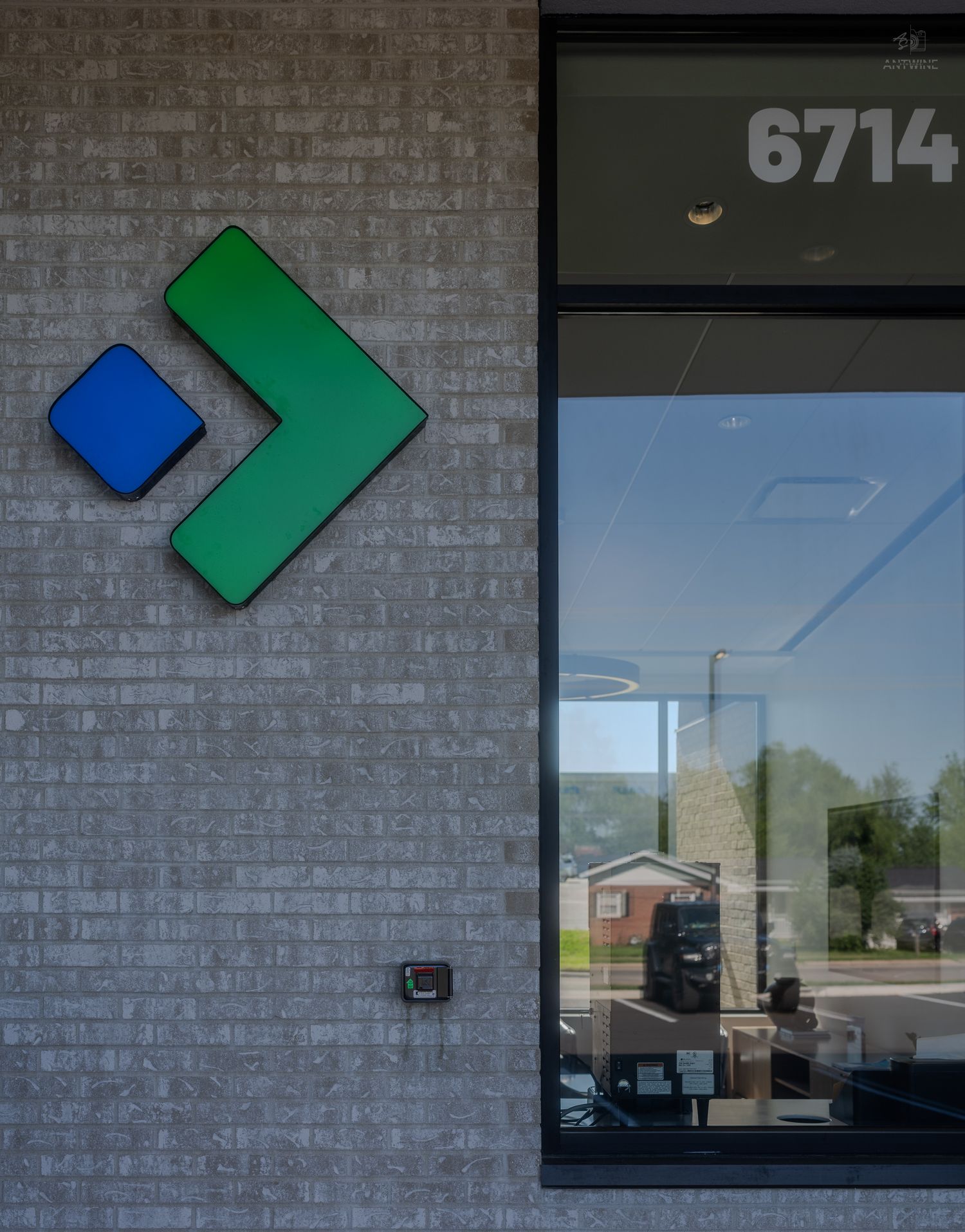 Green and blue arrow logo on brick wall exterior of building number 6714 with glass entrance door.