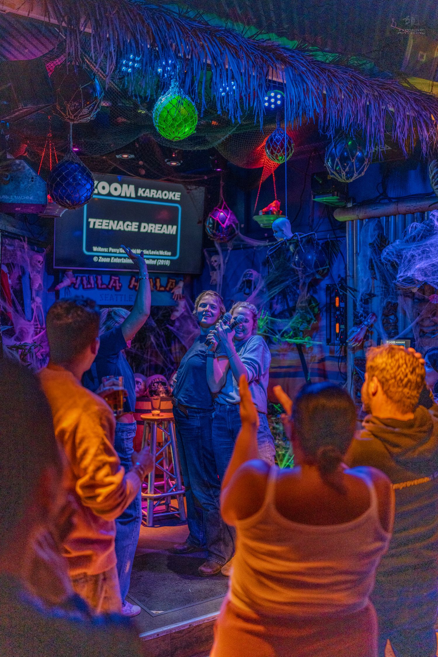 People singing karaoke to Teenage Dream at a colorfully lit tiki bar with Halloween decorations.