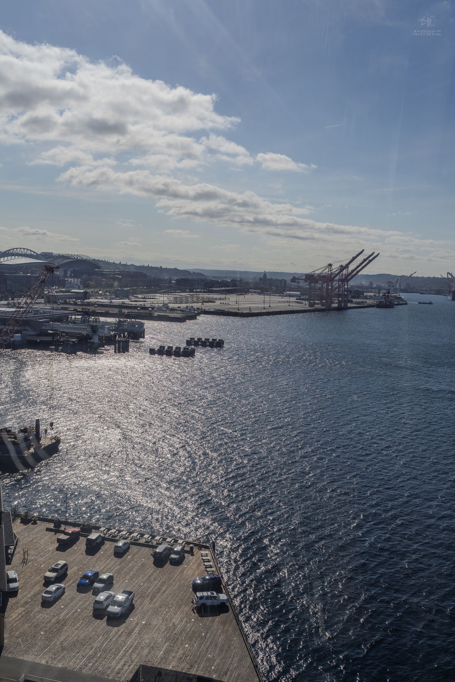 Aerial view of Seattle waterfront port with cranes, docked ships, and sparkling Puget Sound under a partly cloudy sky.