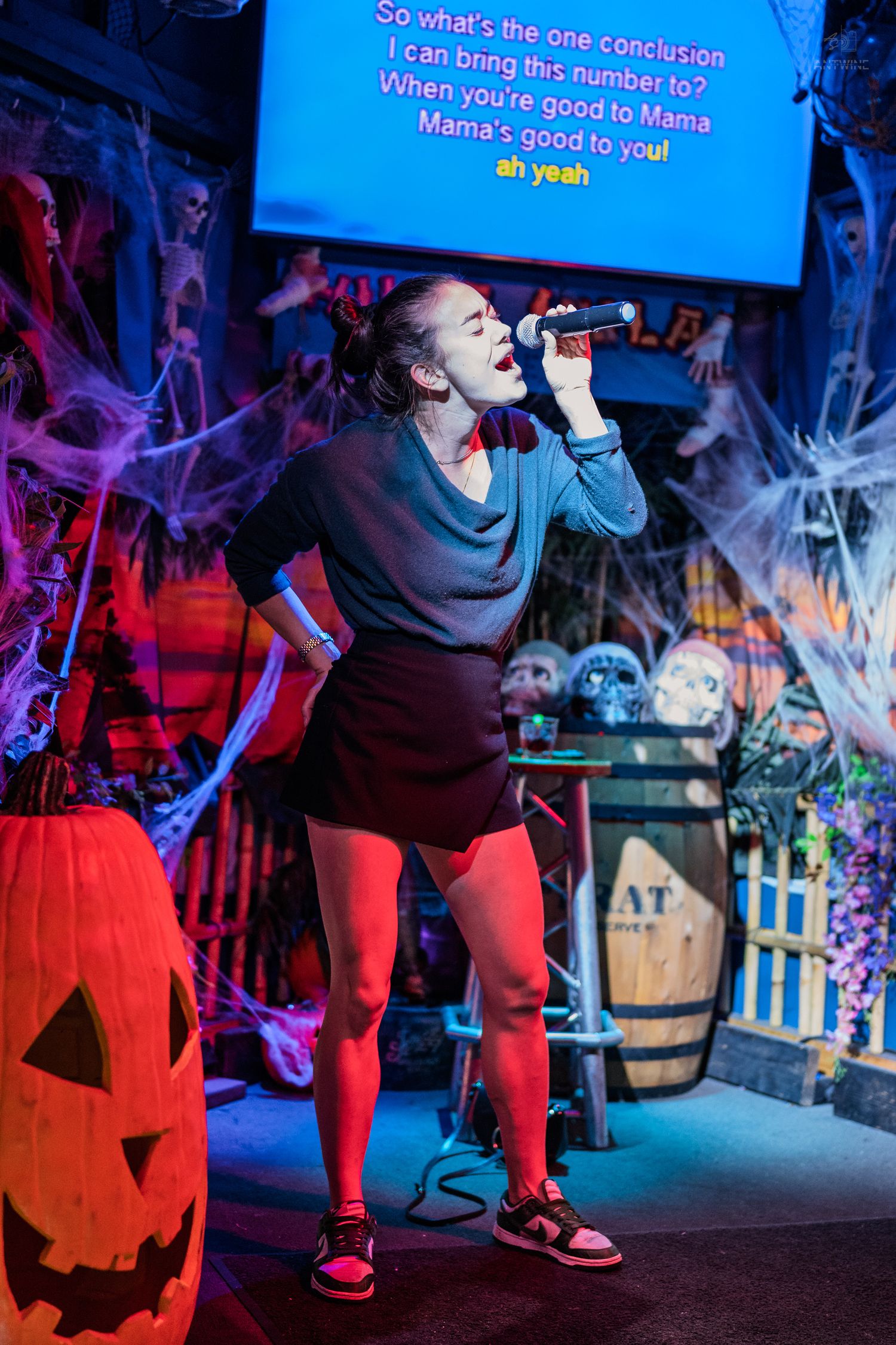 Young woman passionately singing karaoke on a Halloween-decorated stage with spiderwebs and a glowing jack-o-lantern.