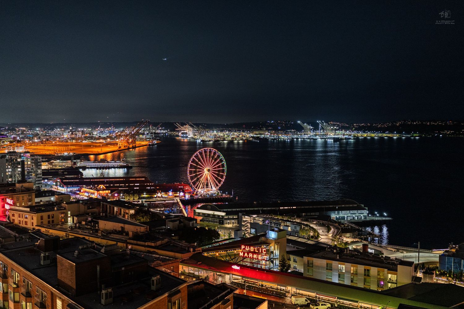 Aerial night view of Seattle waterfront featuring the iconic Great Wheel and city lights reflecting on water.