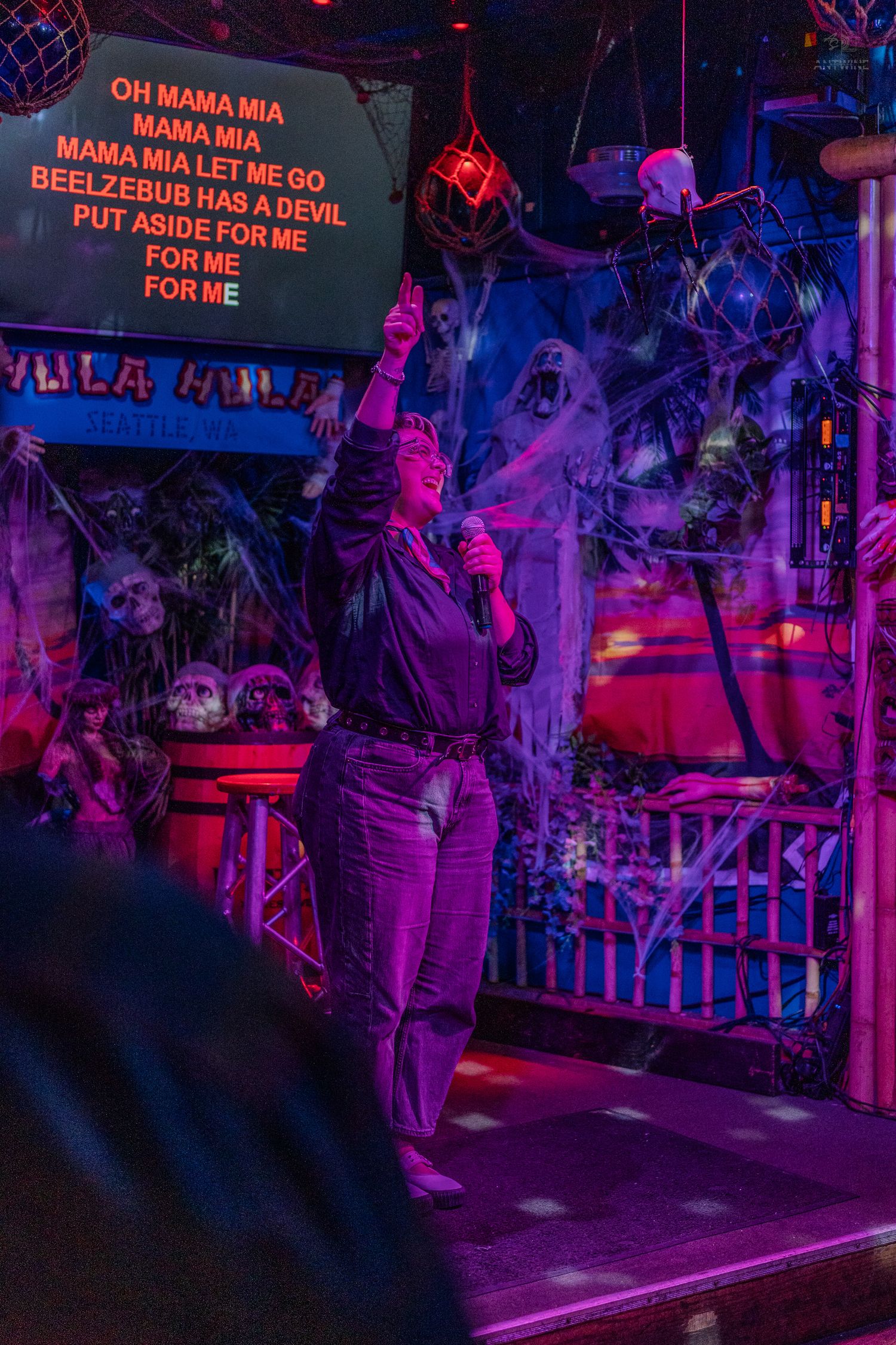 Karaoke performer singing Bohemian Rhapsody under purple neon lights at Halloween-decorated Hula Hula bar Seattle.