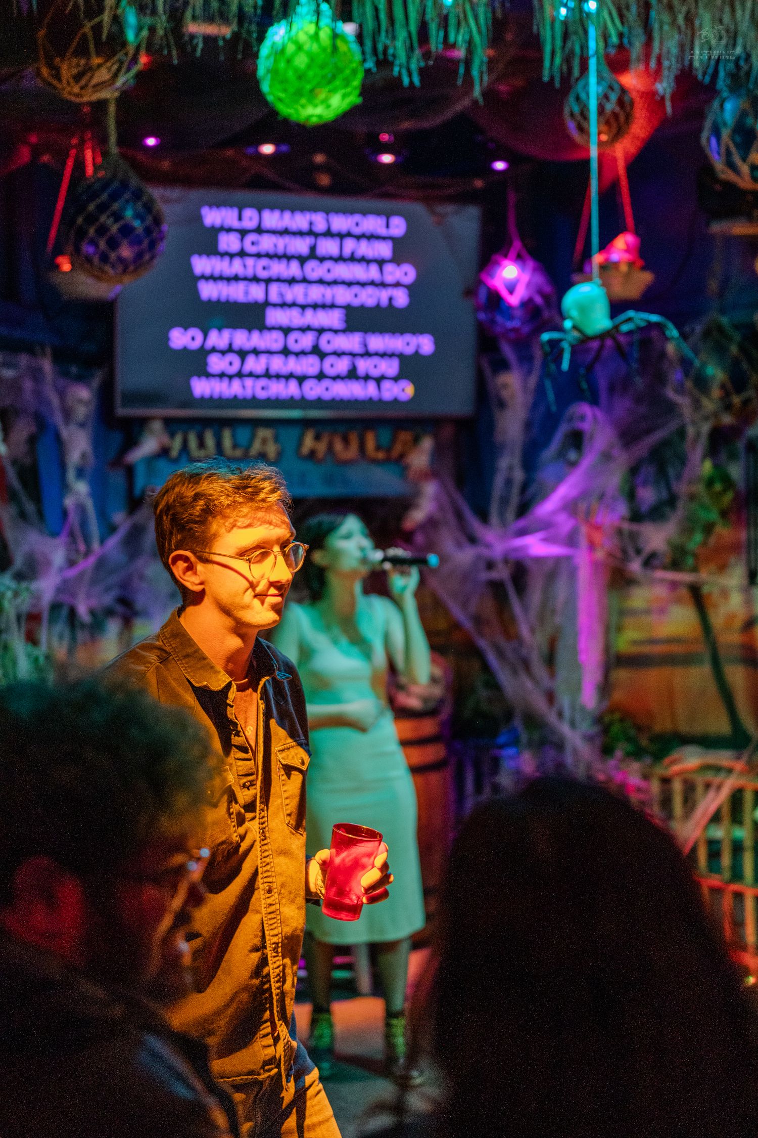 A man holding a red cup at a Halloween-themed karaoke bar with colorful lights and spooky decorations.