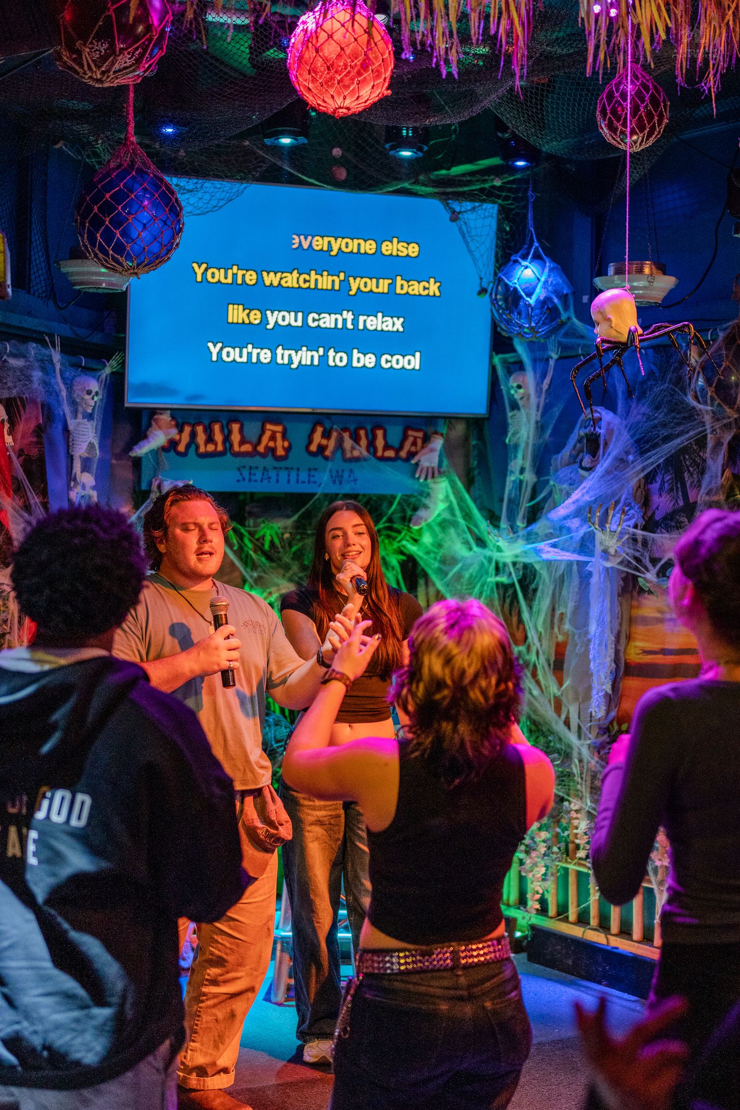 Karaoke performers on stage at Halloween-decorated Hula Hula Seattle bar with cobwebs and neon lights.