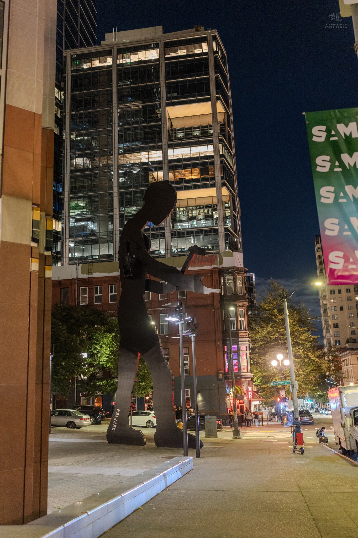 Large metal sculpture of a figure on a scooter illuminated at night in an urban downtown setting.