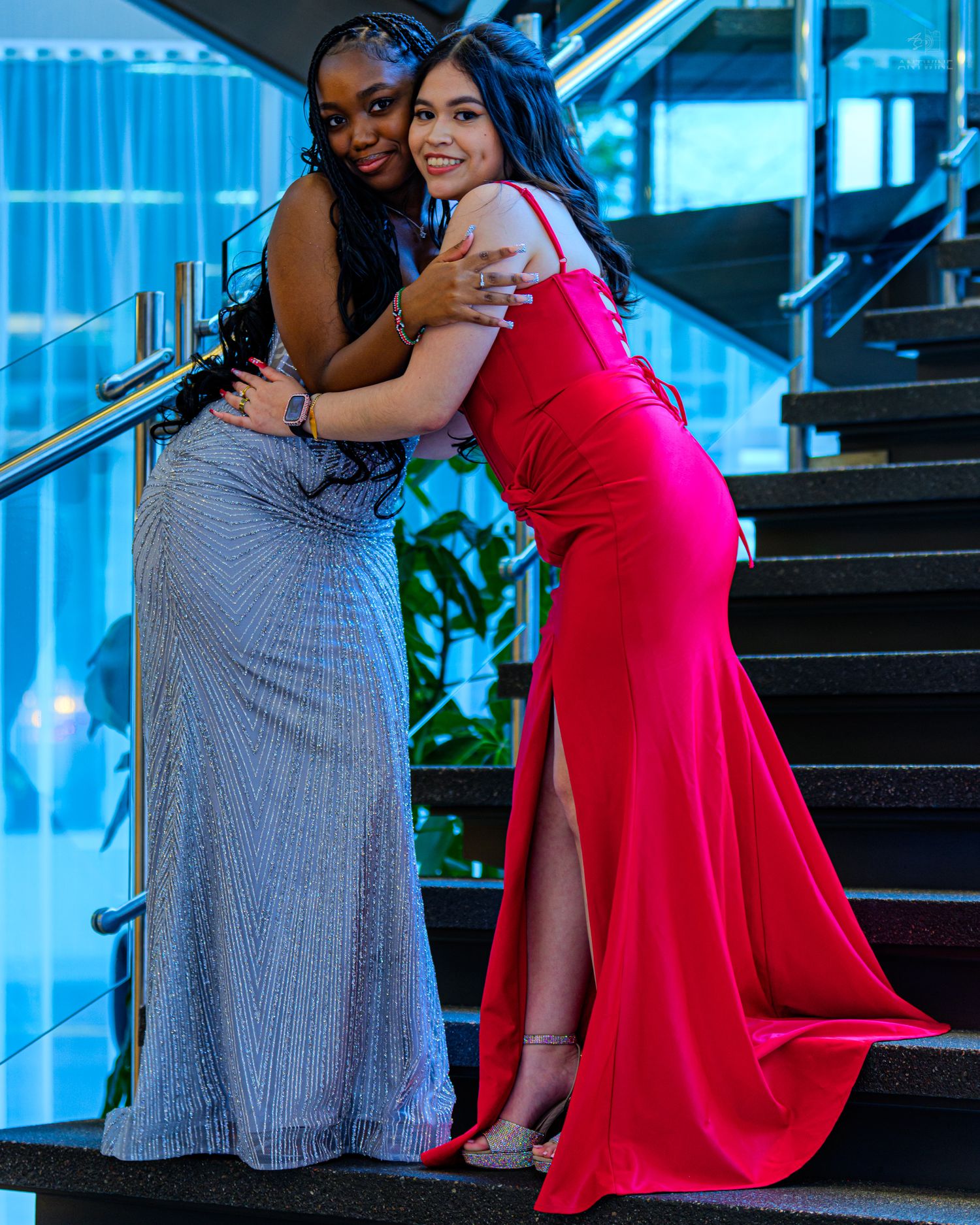 Two young women in elegant formal gowns embracing on stairs, one in silver and one in red dress.
