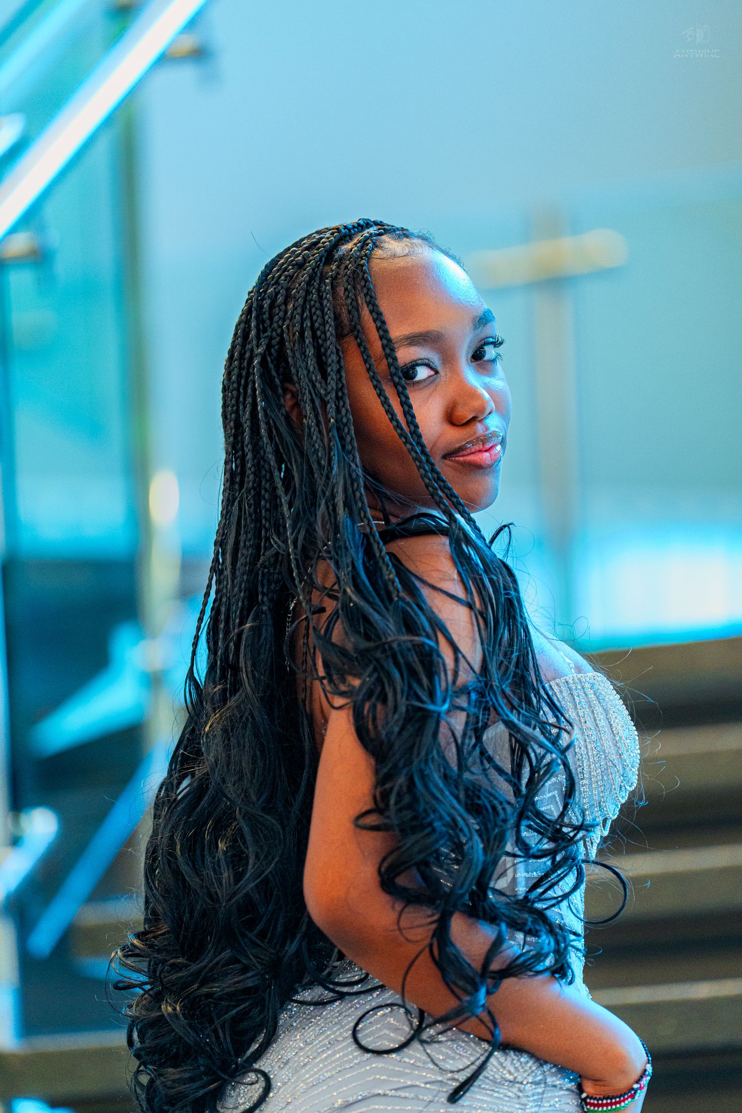 Young Black woman with long box braids poses elegantly in a silver beaded gown near glass staircase.