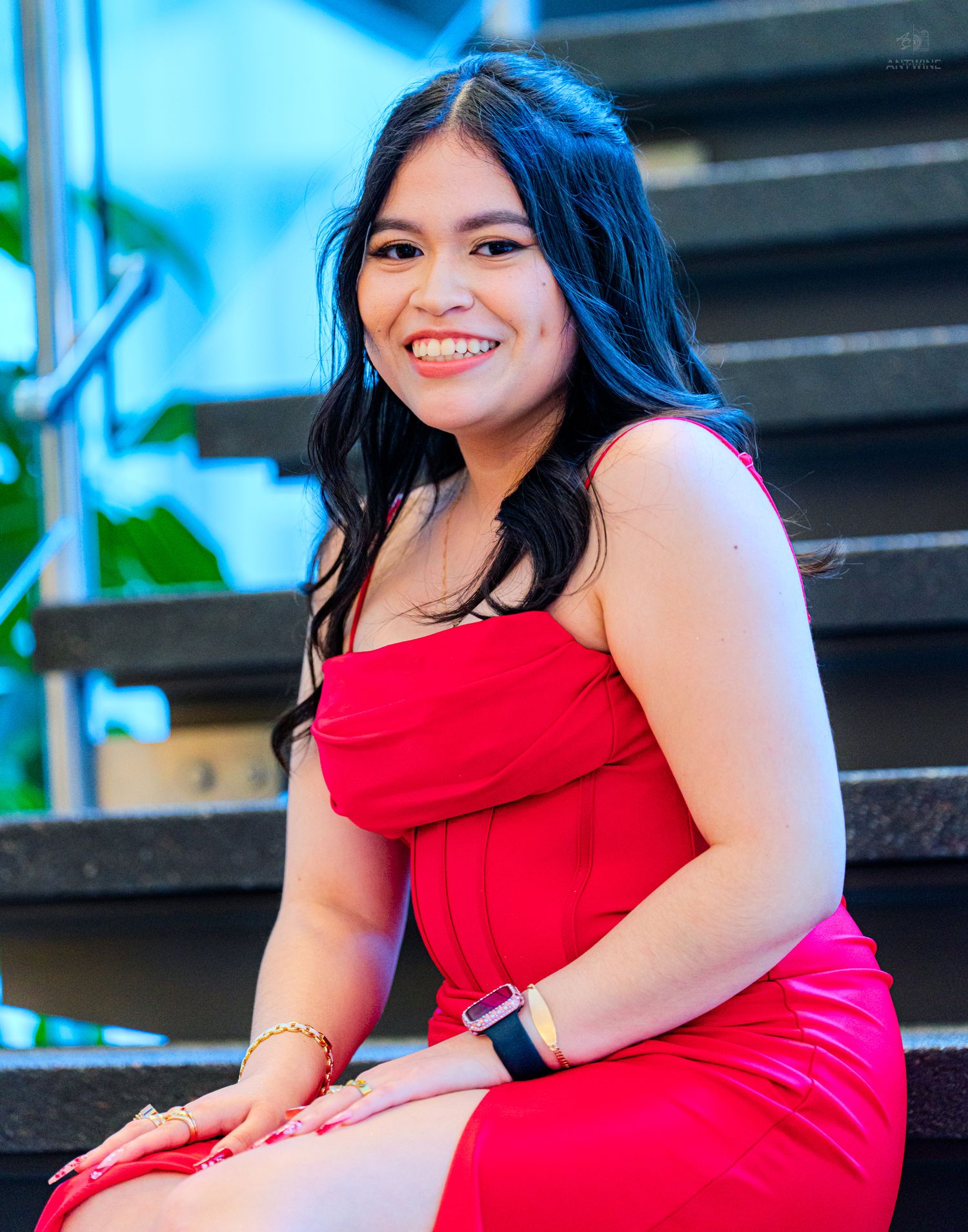 Smiling young woman in red strapless dress seated on stairs with blue-lit background.