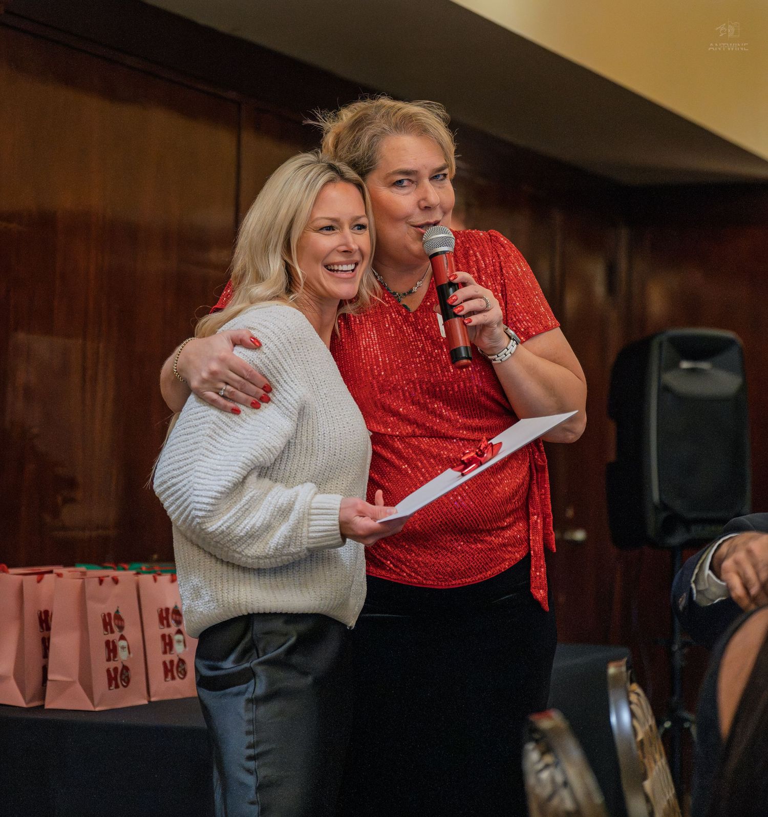 Two women smiling and hugging at a holiday event, one holding a microphone and paper, with gift bags in background.