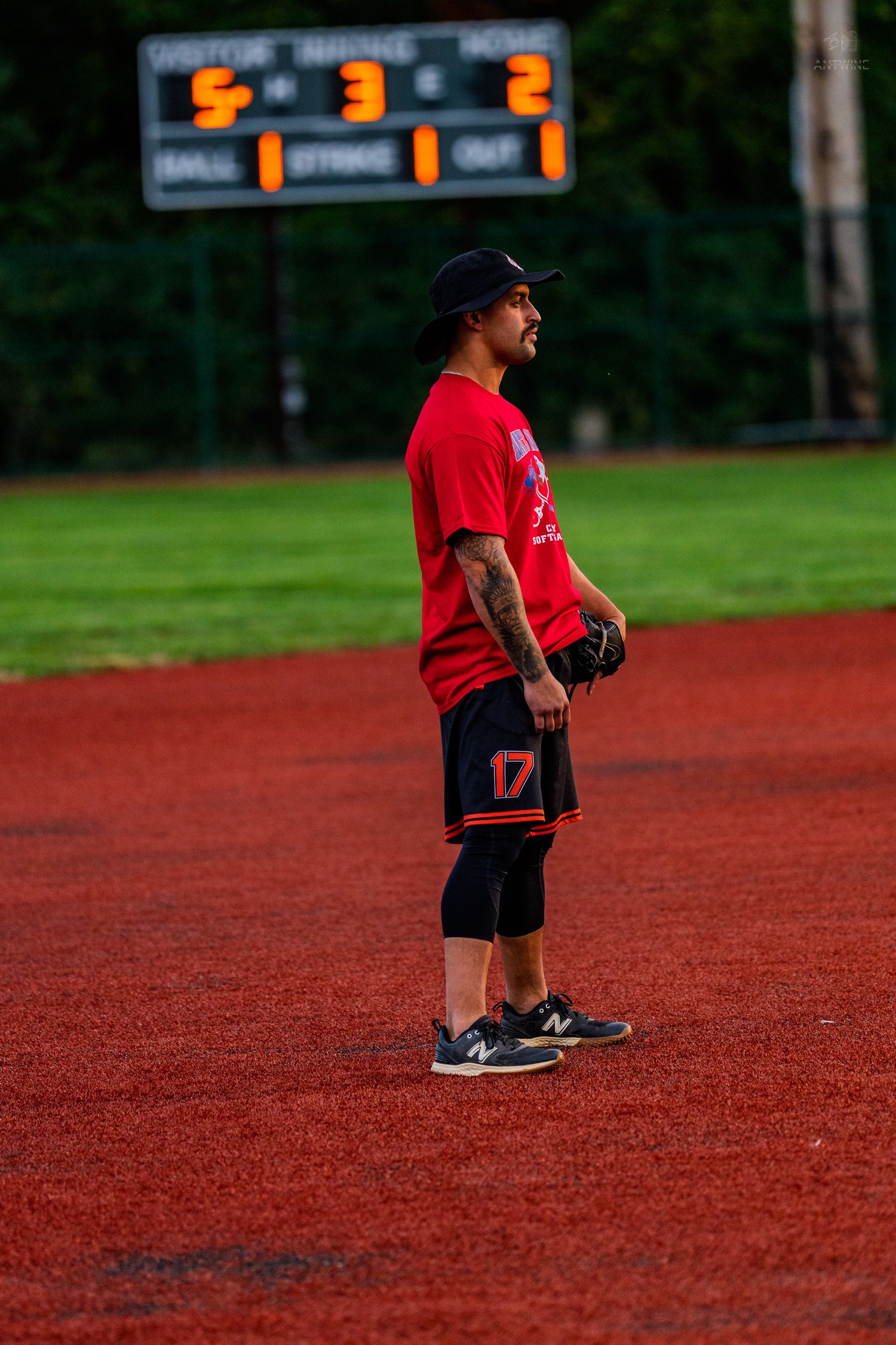 A baseball player wearing number 17 in a red jersey stands on a red dirt infield near the scoreboard.