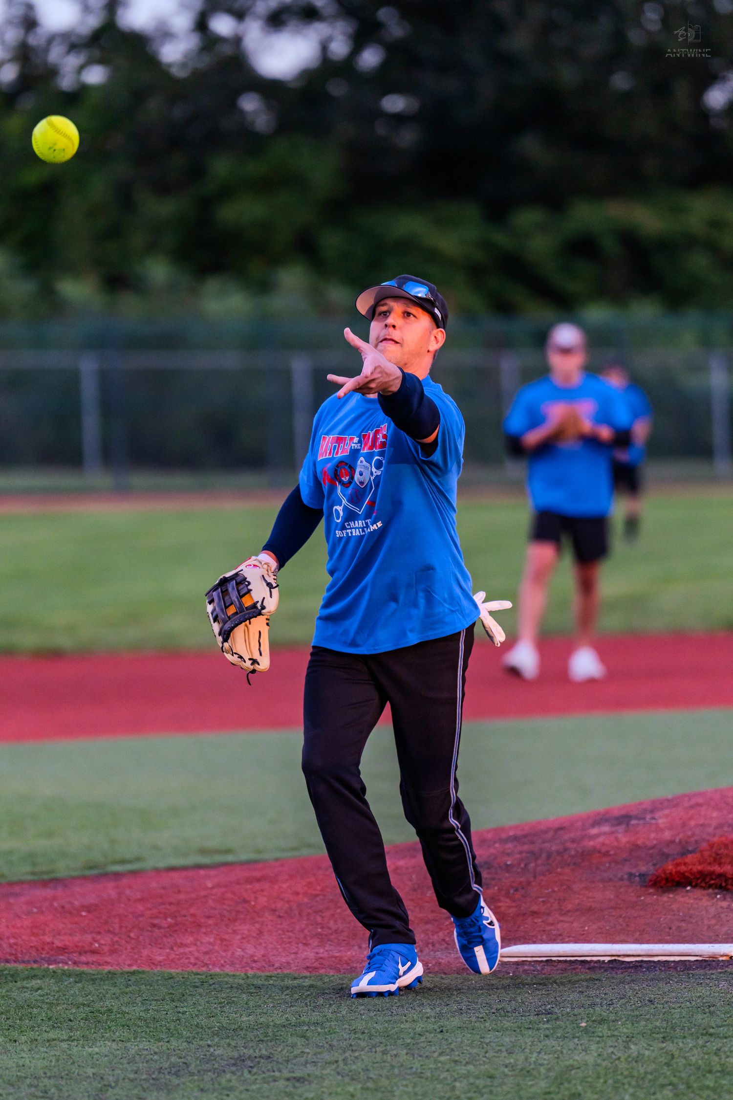 Softball player in blue jersey releases yellow ball mid-throw during evening league game.