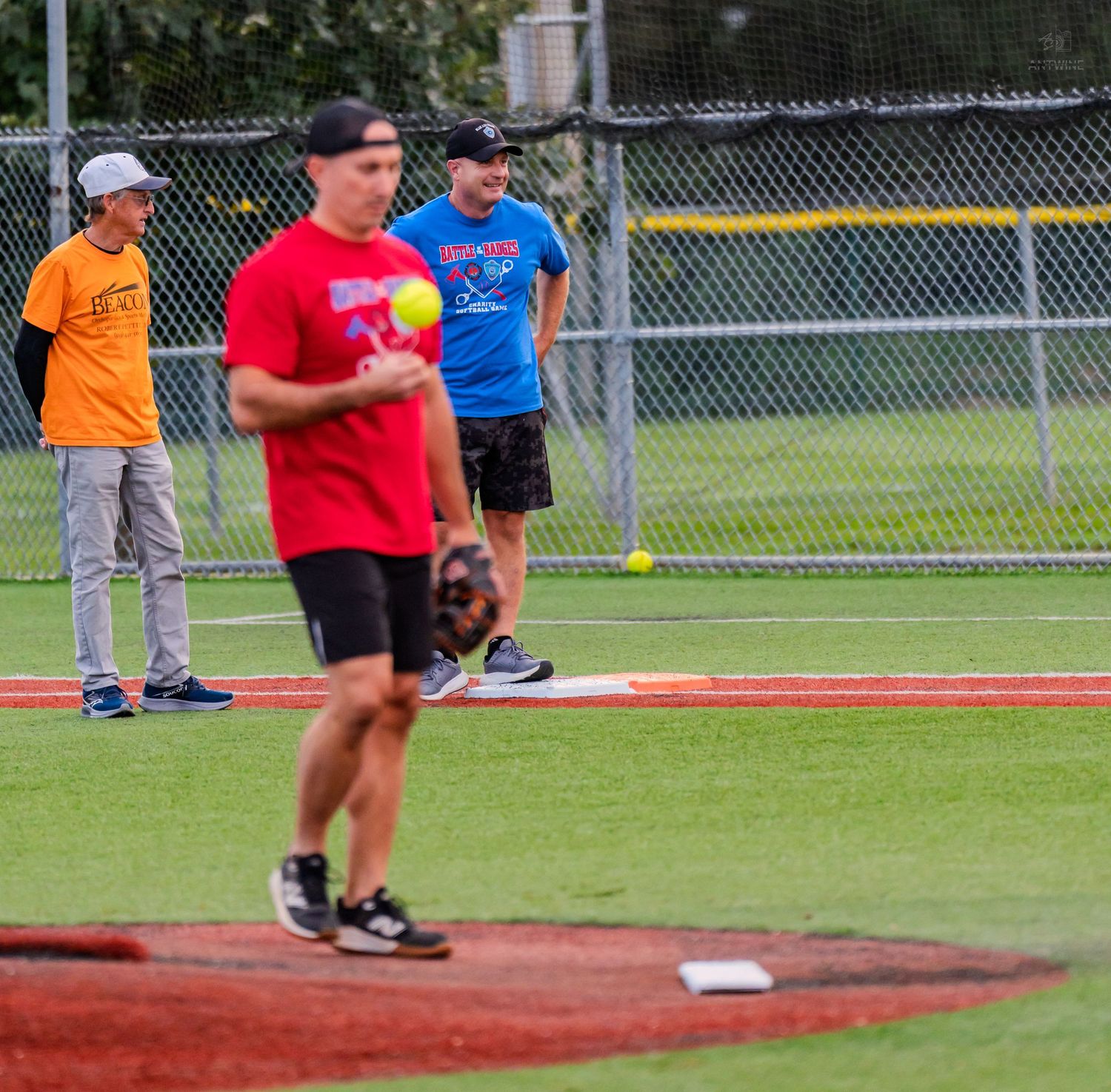 Man in red shirt holds softball on baseball field while players watch during a recreational softball game.