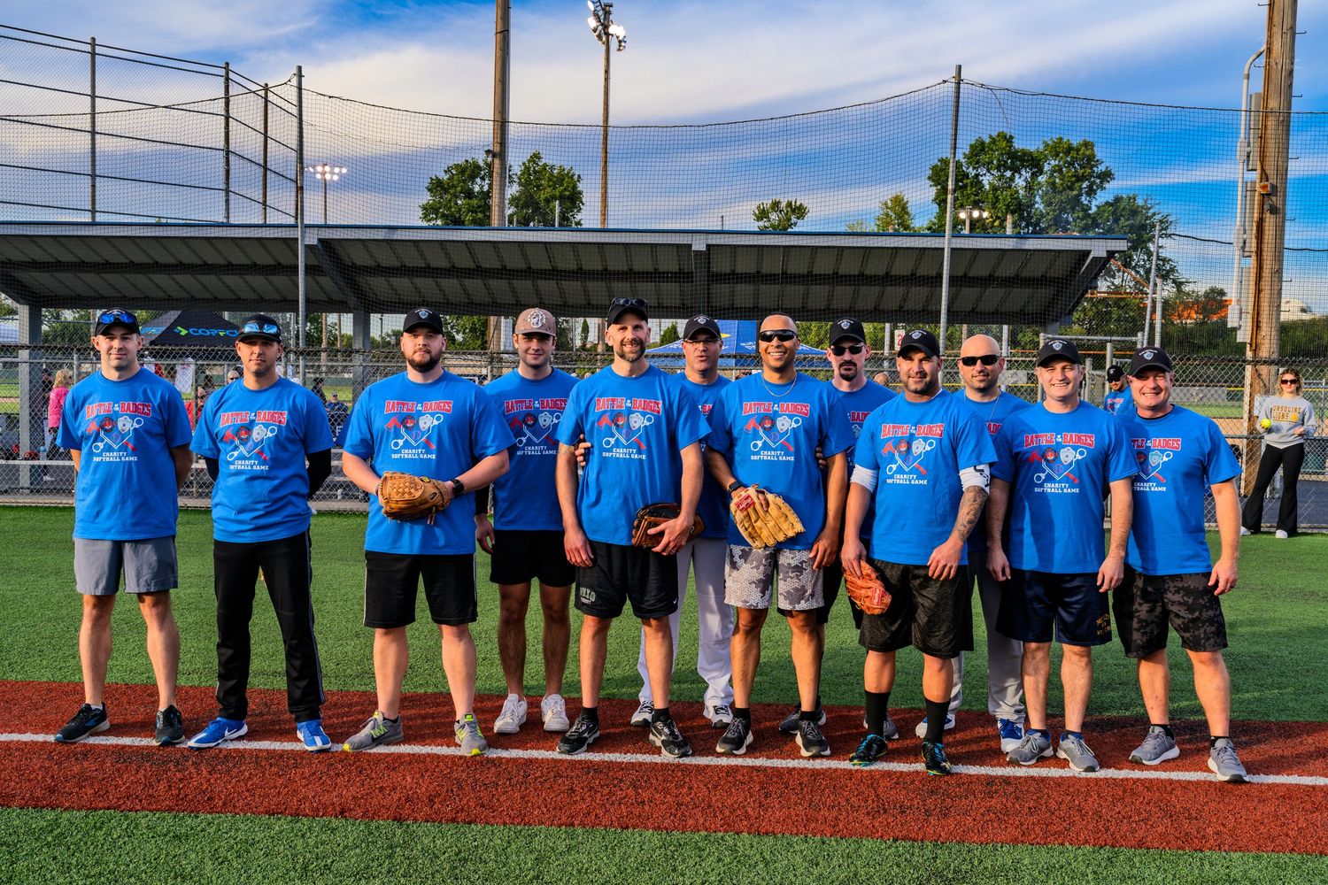 A group of softball players in blue team shirts pose together smiling on a baseball field at dusk.