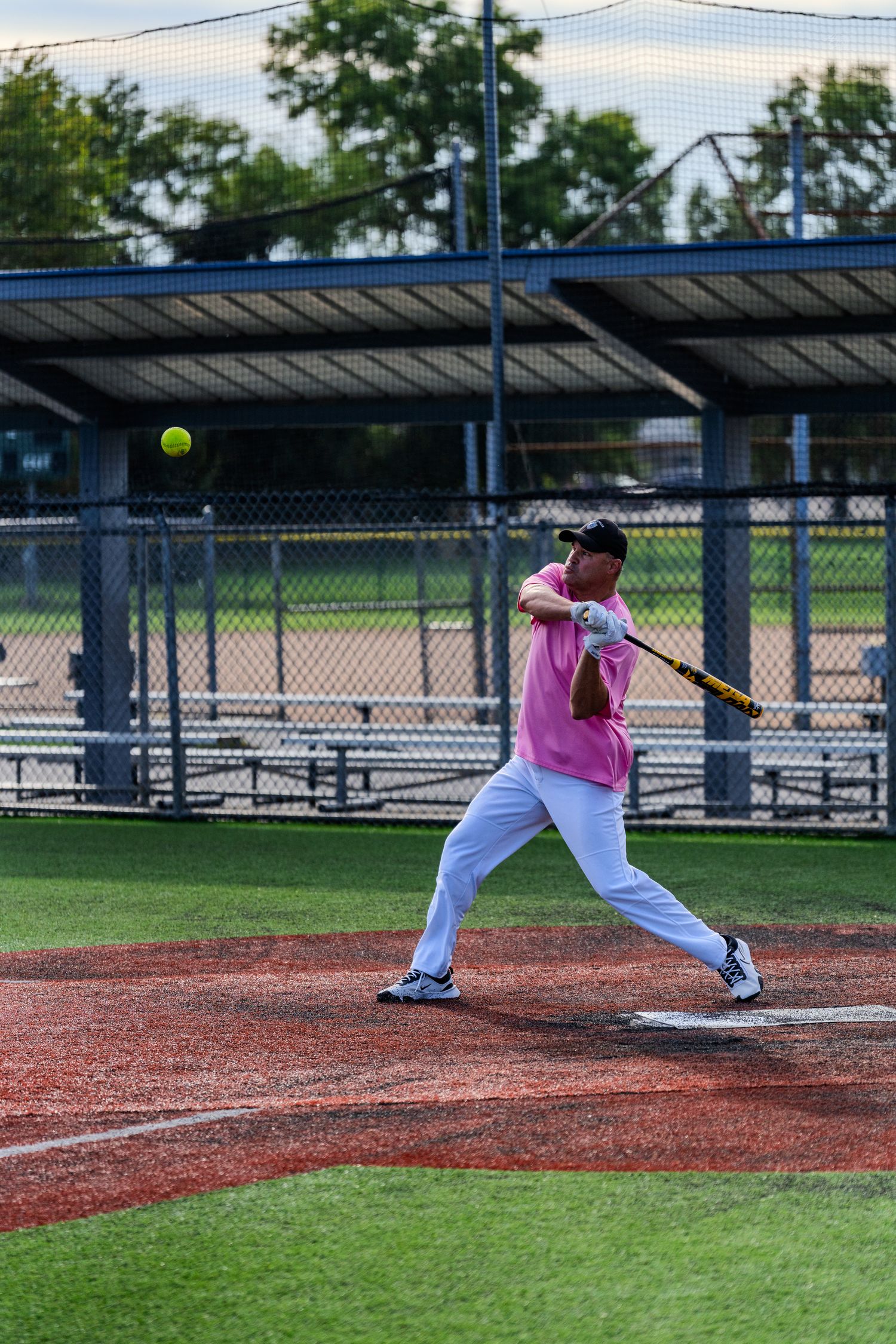 Adult softball player in pink shirt and white pants makes contact with a yellow ball at home plate.