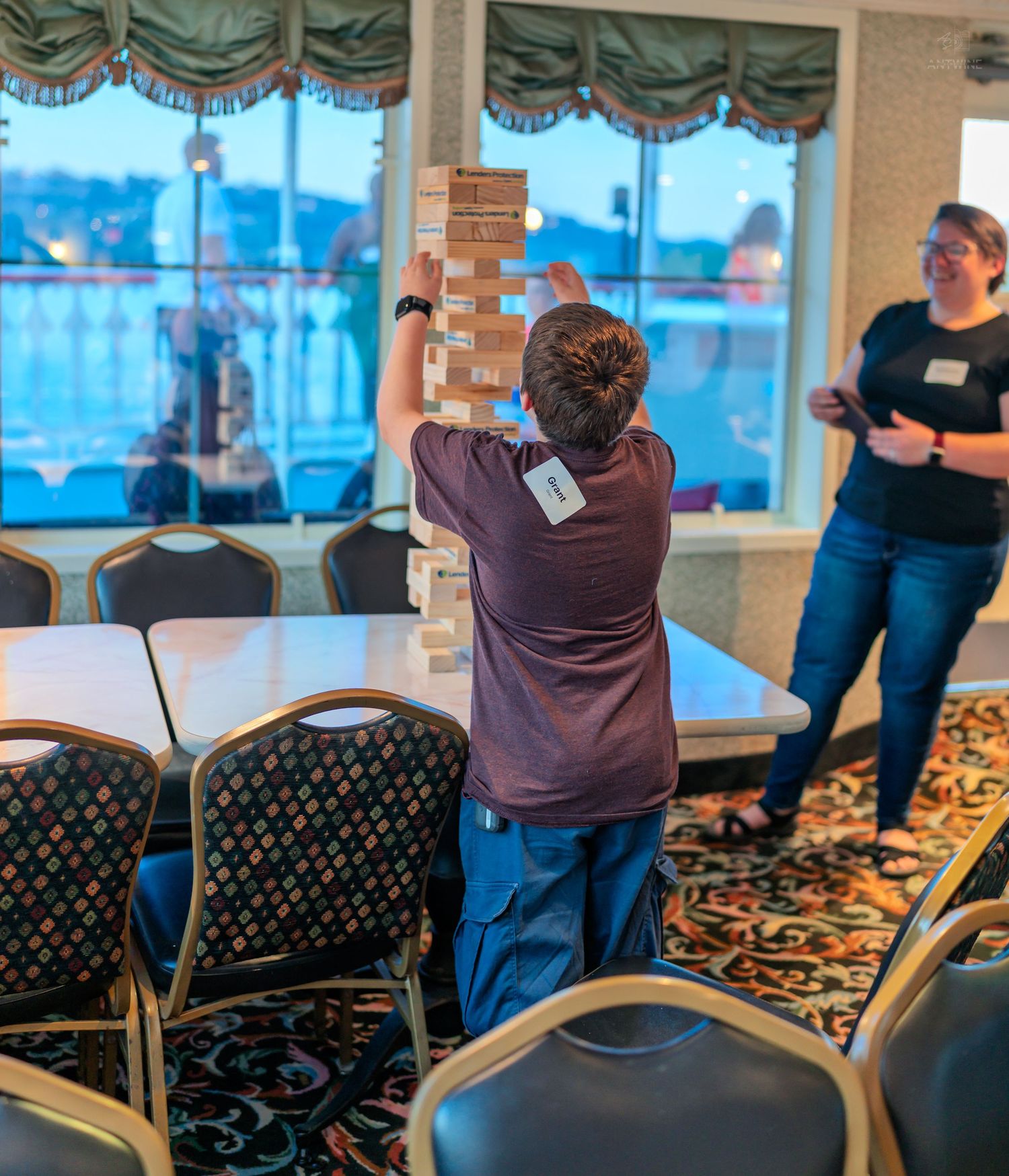 Person carefully balancing a tall Jenga tower during a corporate event in a banquet hall with scenic windows.