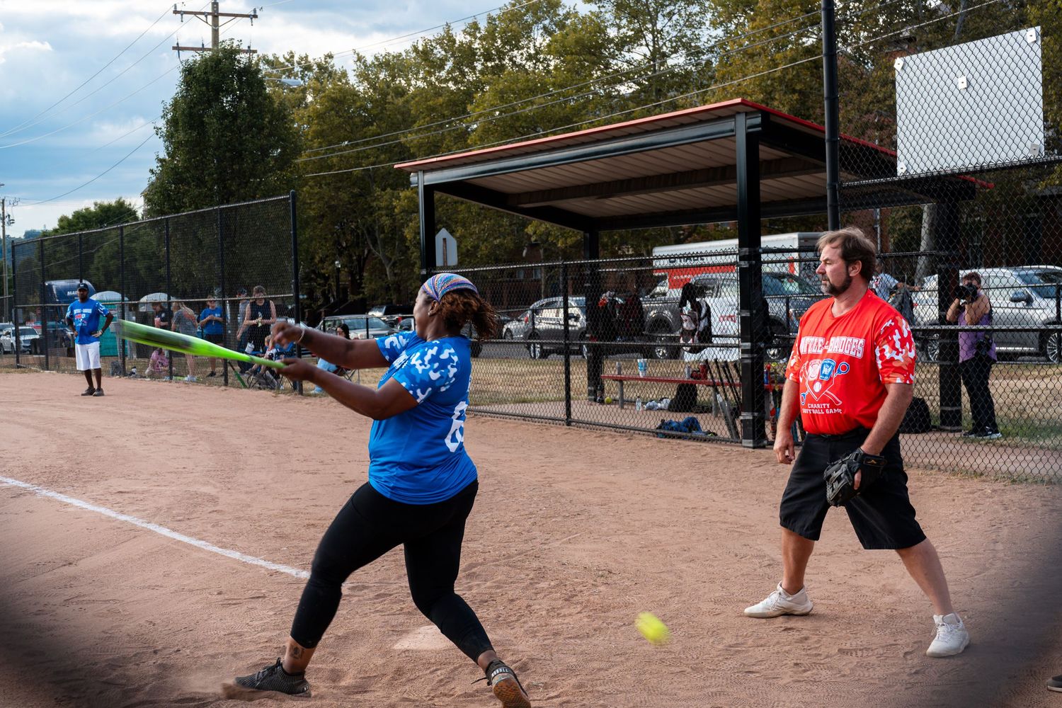 A batter swings at a pitch during a recreational softball game on a dirt field.