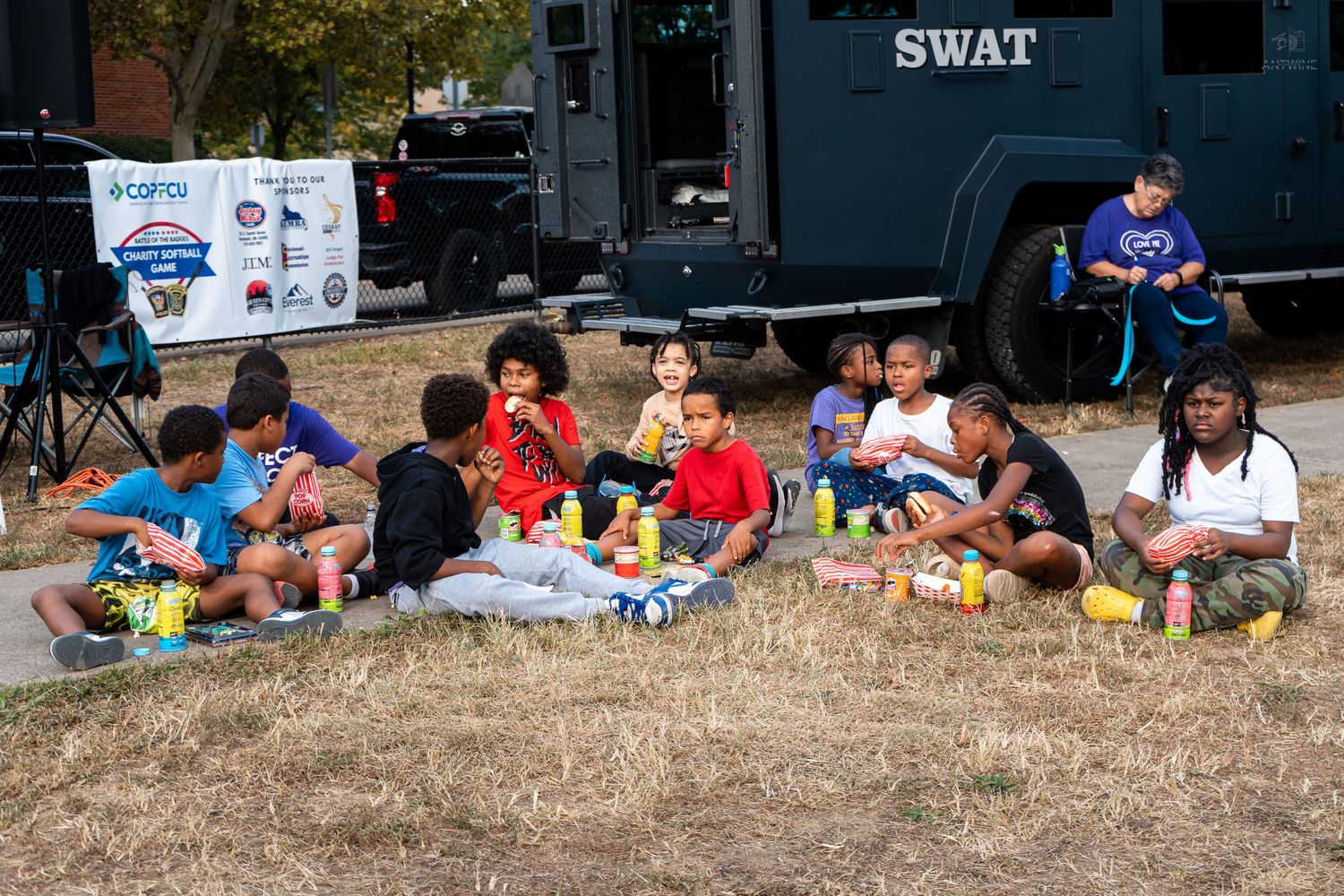 Children sit on grass eating snacks near a SWAT vehicle during a community outreach event on a sunny day.