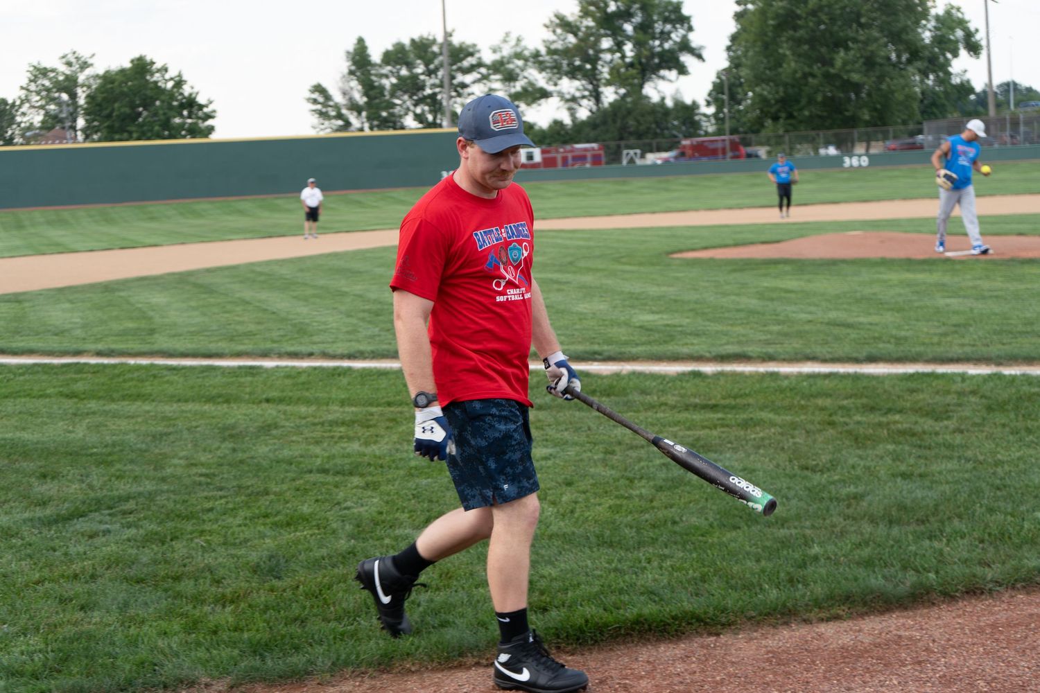 Man in red shirt and baseball cap walks on field holding bat during outdoor baseball practice game.