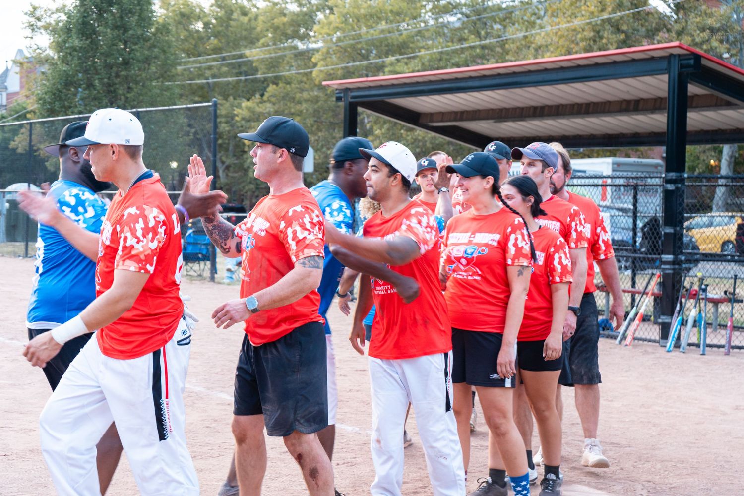 Red and blue team softball players shake hands on a dirt field after a community game outdoors.