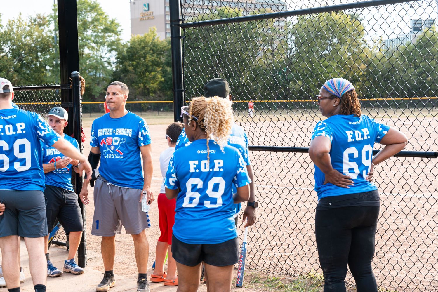 Players in blue FOP 69 jerseys gather along the fence during a community charity softball game.