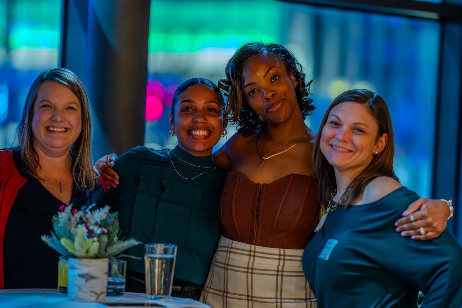 Four women smiling and posing together at a social event with colorful lighting in the background.