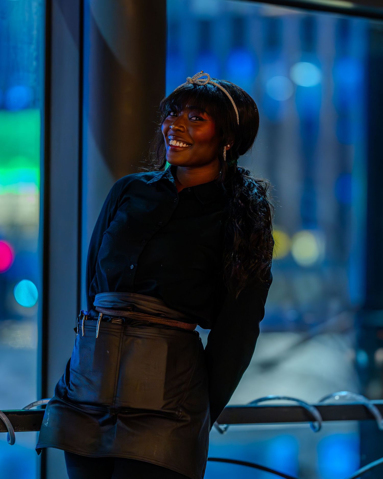 Smiling young Black woman in black outfit and headband posing near city window at night.
