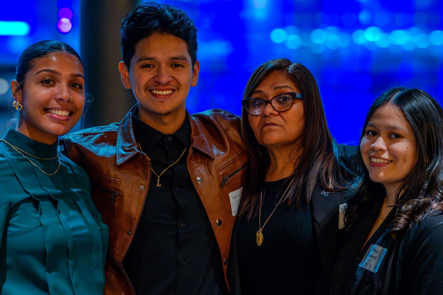 Four young adults smiling together at a blue-lit event, dressed in smart casual attire.