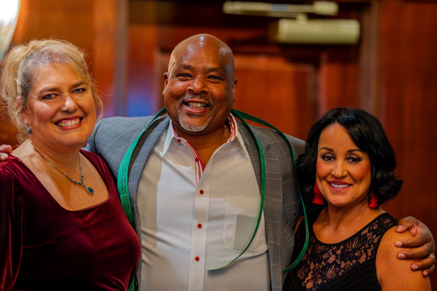 Three smiling friends pose together at a festive event, with two women flanking a man in a gray blazer.