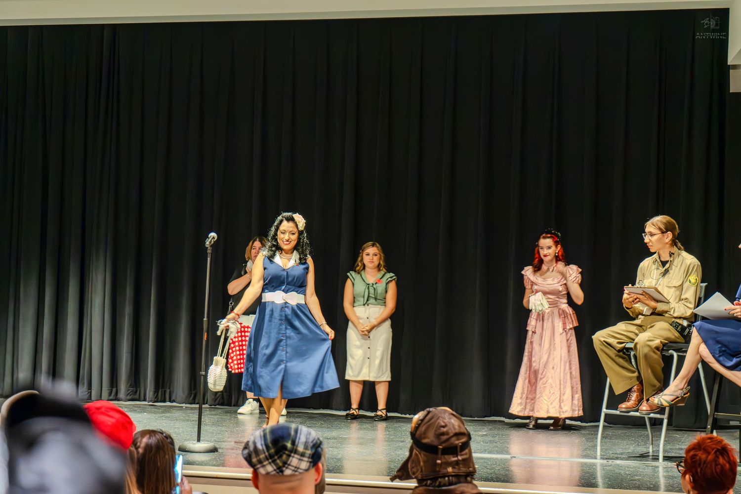 Four performers on stage in costume during a school or community theater production with a microphone stand and dark curtain backdrop.