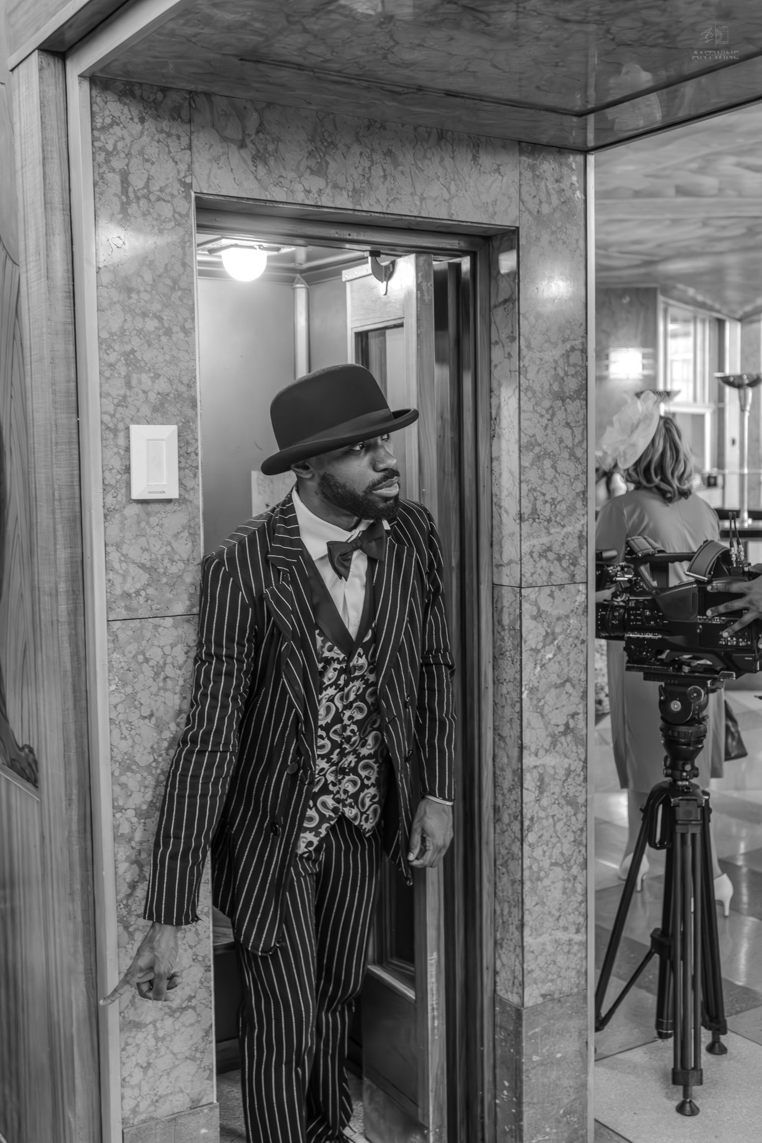 A well-dressed Black man in a pinstripe suit, bow tie, and bowler hat stands in an elevator doorway in a black and white photo.