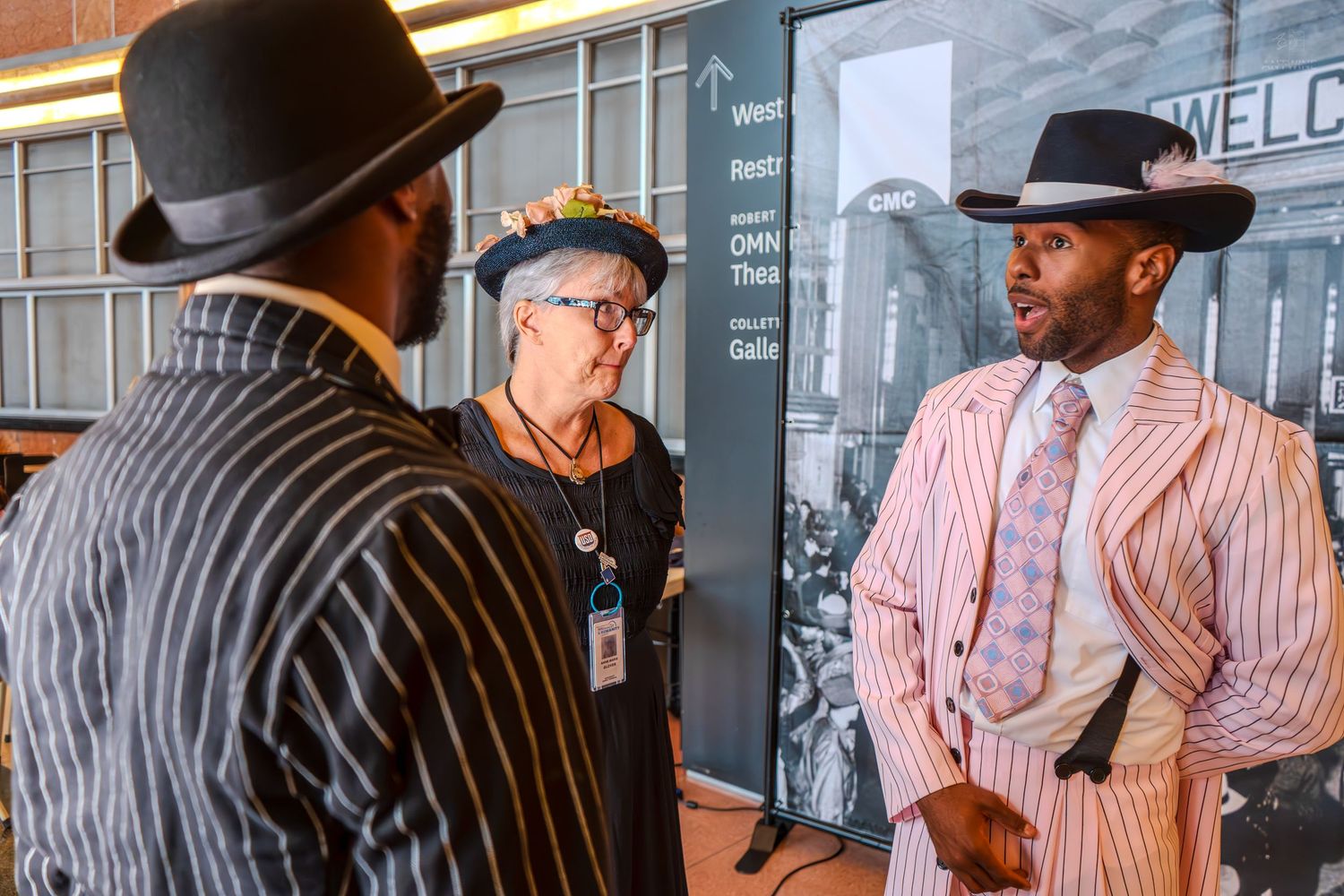 Three people in vintage-style suits and hats converse at a museum exhibit event.
